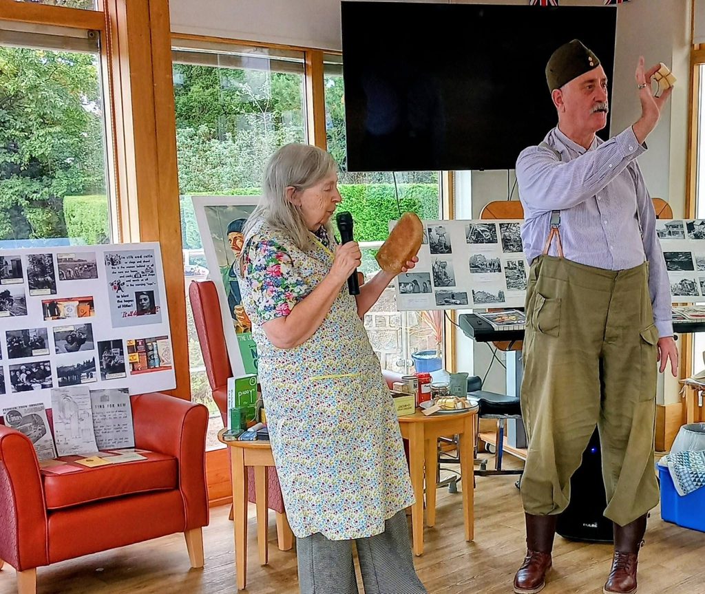 An elderly woman in an apron, sharing Stories and Memories, holds bread and a microphone whilst a man in period dress presents an artefact at a Ler House historical talk with display boards.