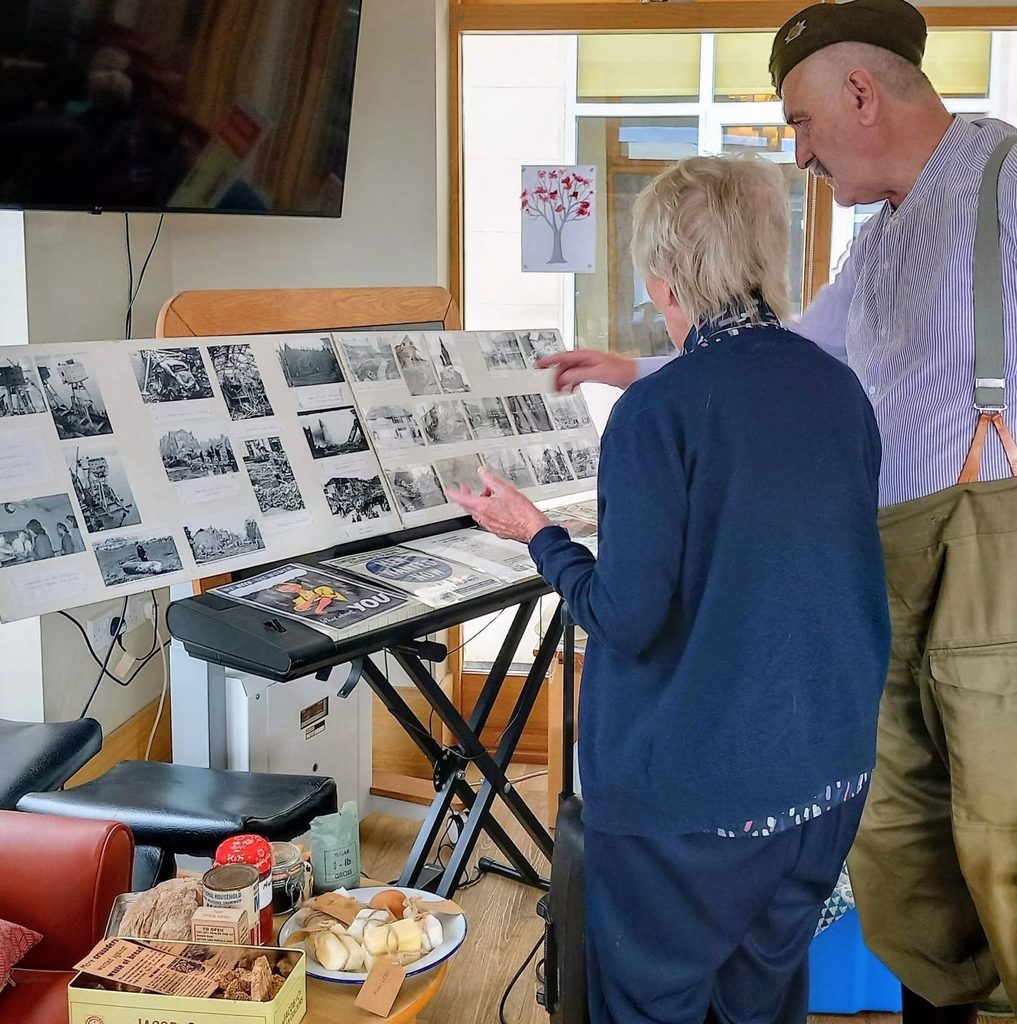 At Lear House, two elderly people stand by a keyboard, reminiscing as they look at black-and-white photographs. Tea and treats are set out nearby in the sunlit room.