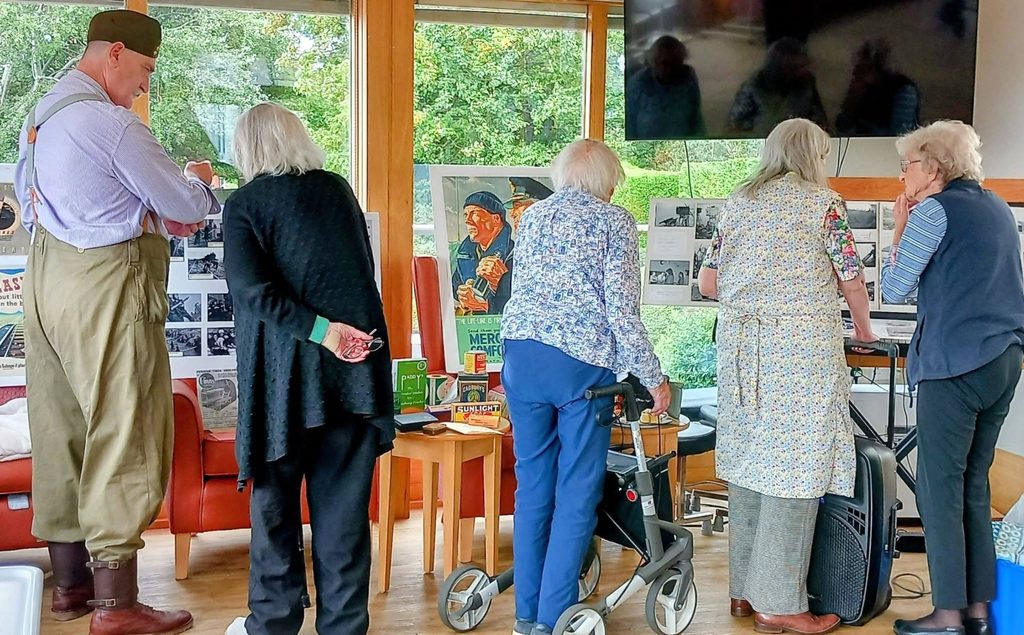 Five older adults, one in fancy dress, look at vintage Heswall Home Guard posters and photos at Lear House. Tables hold memorabilia as they reminisce; some have mobility aids. Large windows show greenery outside.