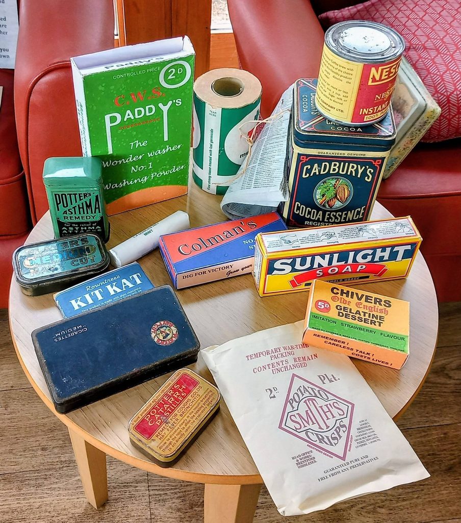 A round wooden table at Lear House showcases vintage product packaging, conjuring stories and memories, with red chairs in the background—possibly once assembled by the Heswall Home Guard.