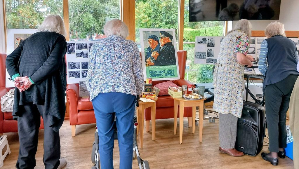 Four elderly women at Lear House look at Merchant Navy memorabilia, reminiscing and sharing stories in a bright room with large windows, red chairs, and references to the Heswall Home Guard.