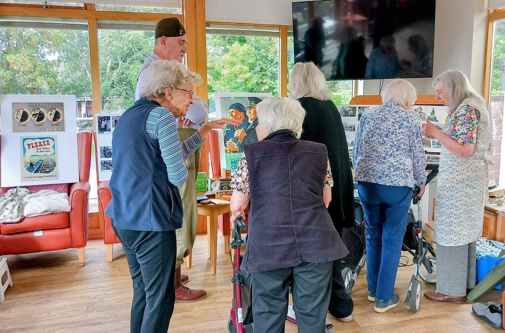 A group of elderly people and a man in a cap share stories and memories in a bright room at Lear House, adorned with Heswall Home Guard posters. Some use walking frames, and there is a large telly on the wall.