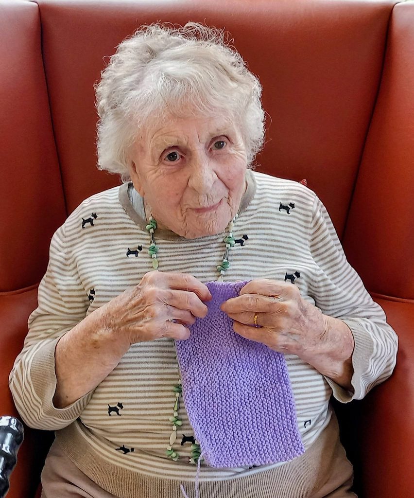An elderly woman with white hair sits in a red chair, wearing a striped top with dog motifs and a beaded necklace, her needles clicking as she enjoys her winter project, knitting a purple piece of fabric.