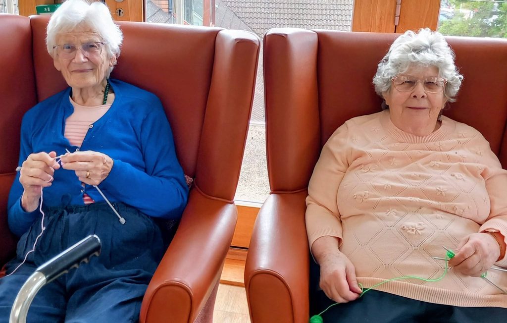 Two elderly women sit in brown armchairs, knitting winter projects. With wool and needles in hand, they enjoy a peaceful Knit and Natter indoors; a walking frame is partly visible in the foreground.