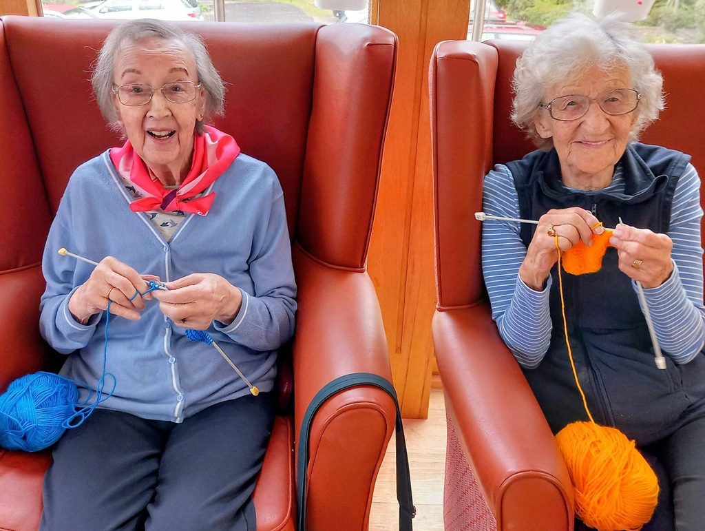 Two elderly women sit in red armchairs, smiling and knitting for their winter project. Needles clicking, they enjoy a jolly Knit and Natter with blue and orange wool balls resting on their laps.