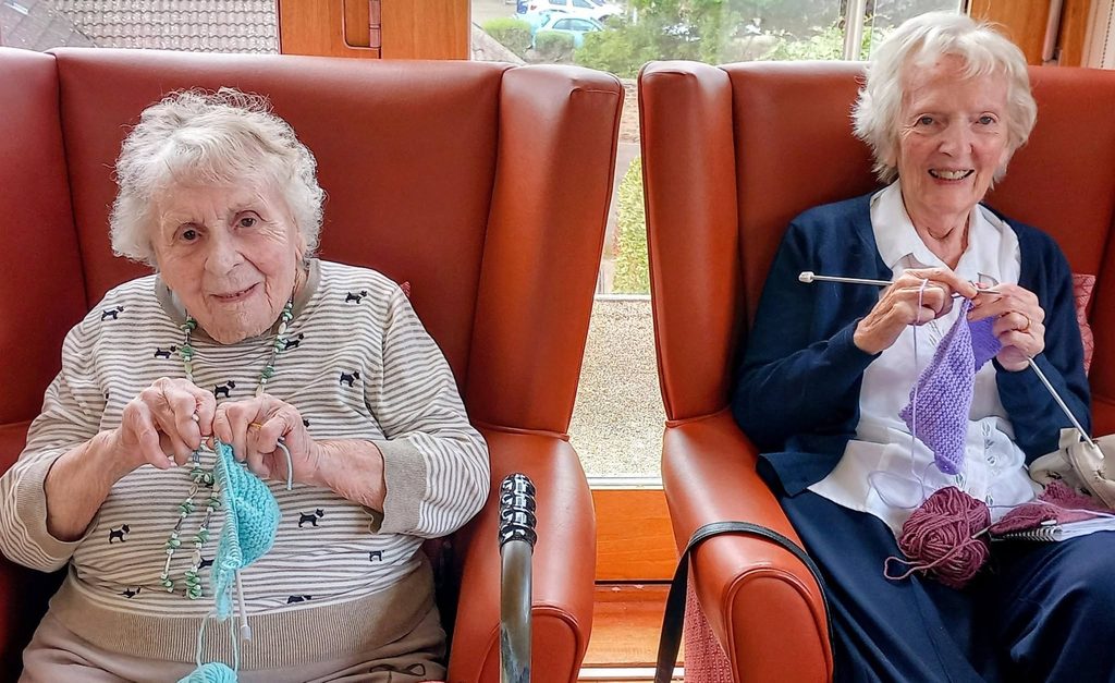 Two elderly women sit in armchairs, smiling and knitting a cosy winter project with colourful wool and needles. A window behind them shows a tranquil outdoor scene.