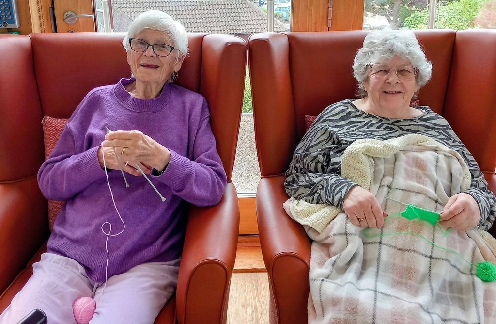 Two elderly women share a Knit and Natter session: one knits with pink wool, the other, wrapped in a checked blanket, holds green wool. Both smile in a bright room with windows—a lovely winter project setting.