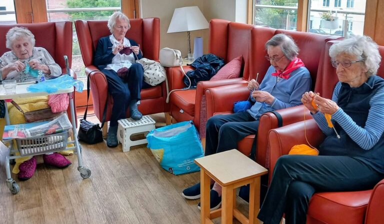Four elderly women sit in armchairs in a light-filled room, knitting and chatting about their latest winter project. A trolley with magazines and drinks is nearby, shopping bags on the floor, creating a calm, relaxed atmosphere.
