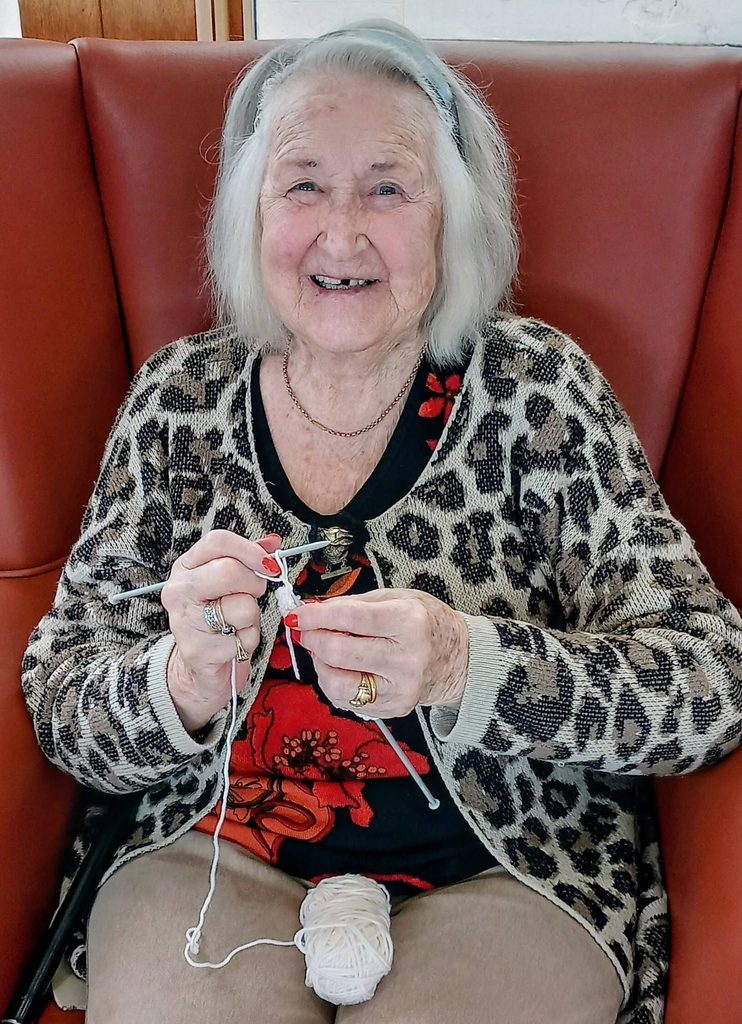 Elderly woman with grey hair sits on a red chair, smiling as she knits a cosy winter project with white wool, ideal for her next Knit and Natter gathering.