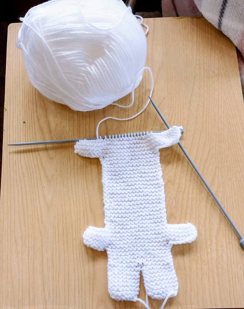 A half-knitted white teddy bear shape on needles lies on a wooden surface, with a ball of white wool nearby—a lovely winter project for your next Knit and Natter get-together.