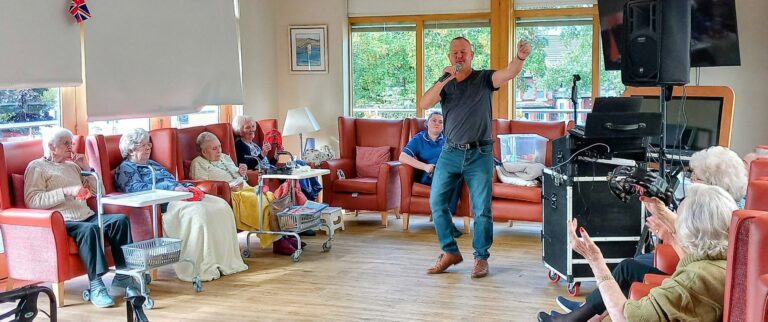 A man sings into a microphone as elderly people sit on chairs and sofas, some applauding, in a brightly lit room with large windows and an audio system.