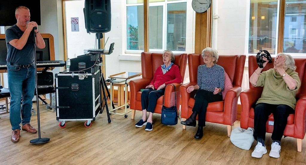 Mark Birch sings into a microphone at Lear House as three elderly women enjoy the performance—one applauding, another smiling, and the third playing a tambourine. Sound equipment and a keyboard are positioned behind them.