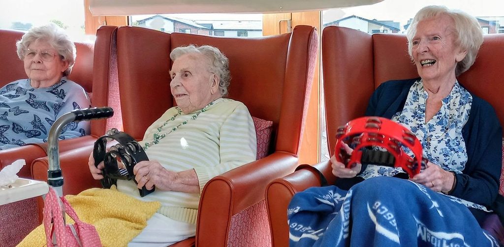 Three elderly women sit in armchairs; two play tambourines. One has a walking frame. All wear spectacles, and one laughs as they reminisce indoors, with blankets across their laps.
