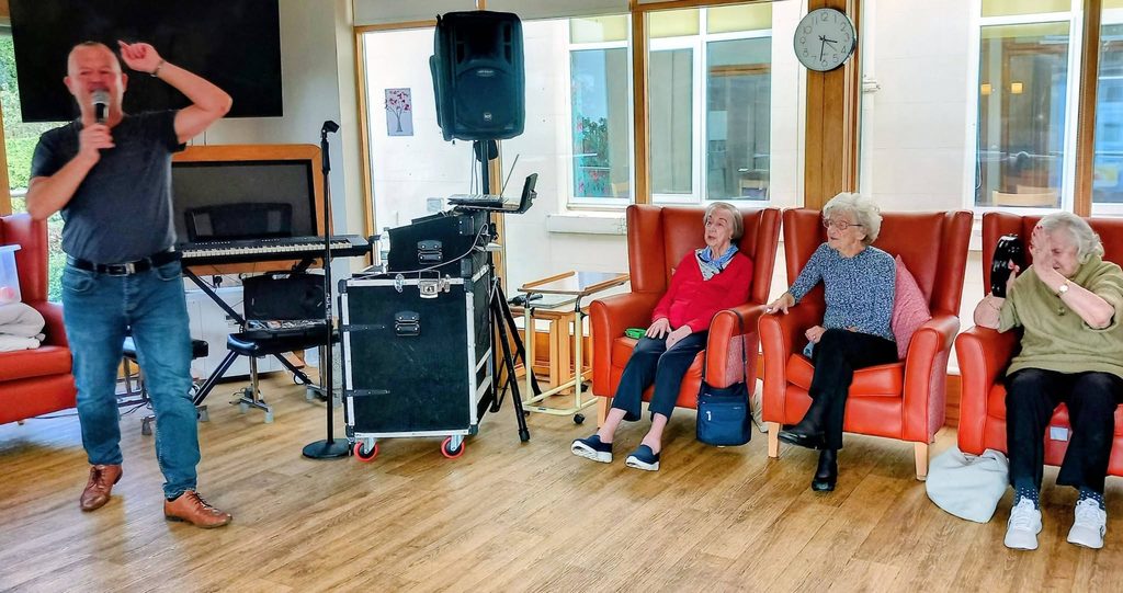 Mark Birch sings into a microphone at Lear House, whilst three older women sit in orange armchairs, smiling and enjoying the music in a brightly lit room with musical equipment and a clock on the wall.
