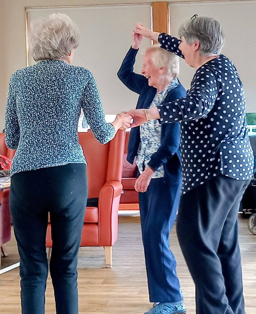 Three elderly women make memories as they dance hand in hand in a bright room. One smiles to the music, while orange chairs and a wheelchair rest quietly in the background.