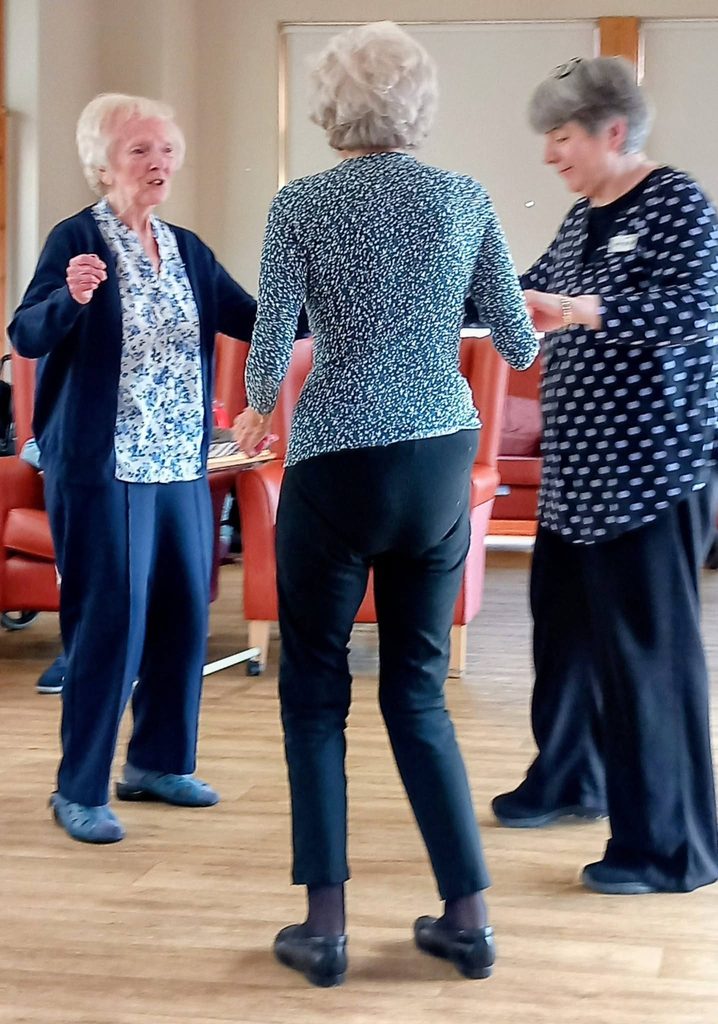 Three older women stand holding hands in a circle at Lear House, seemingly dancing or moving to music in a room with wooden flooring and red chairs.