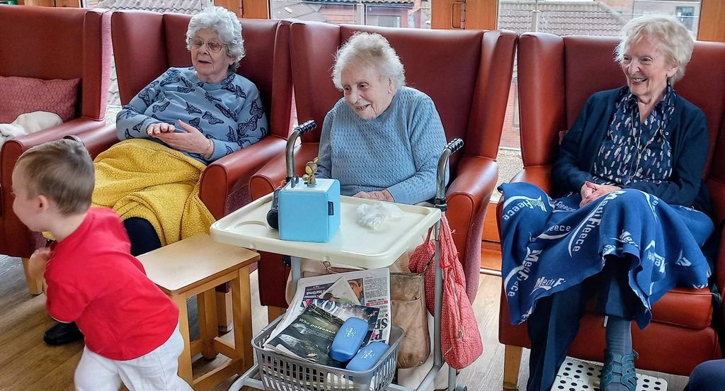 At Lear House Home Nursery, three elderly women relax in armchairs while a young child in a red top and white trousers plays nearby. A tray with items and magazines sits between two of the women.