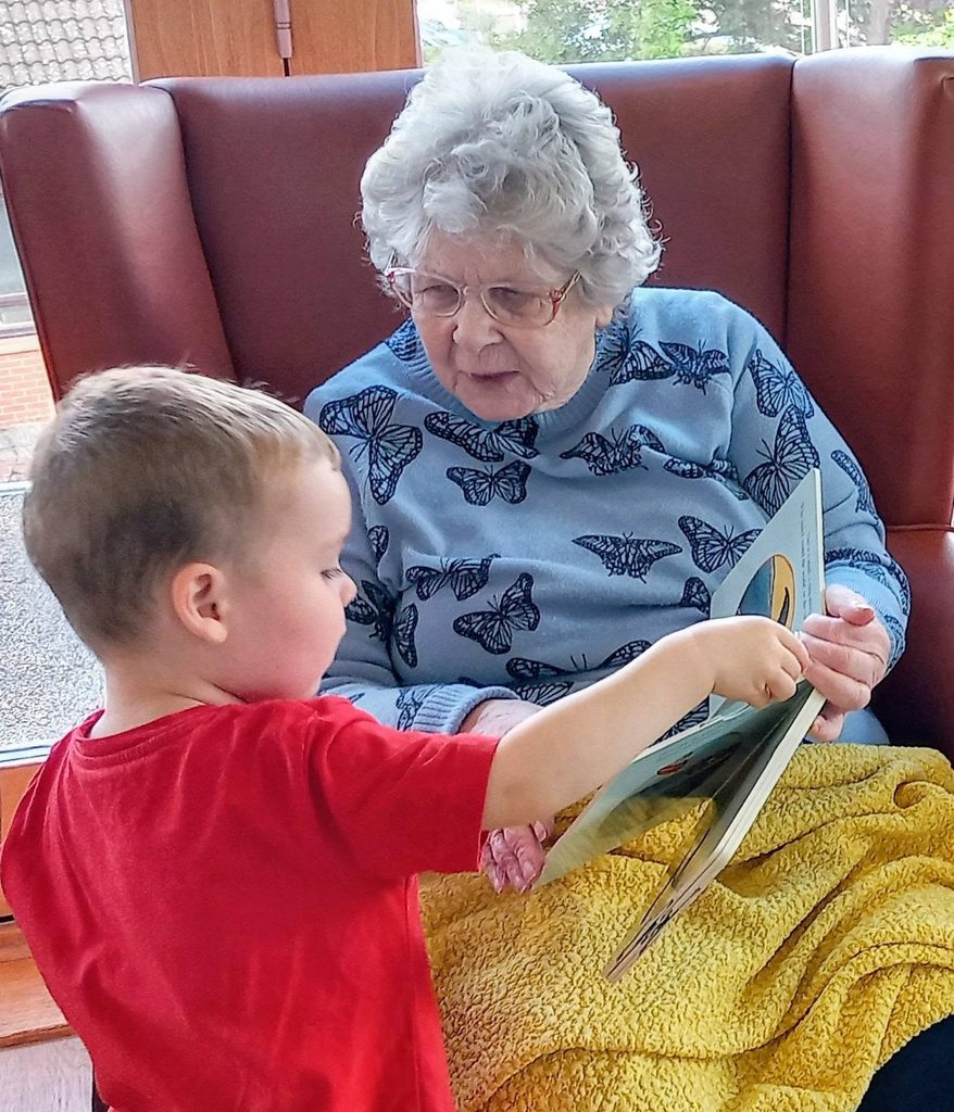An elderly woman with grey hair and glasses sits in a chair draped with a yellow blanket at Lear House, reading a book with a young boy in a red jumper who is pointing at the book.