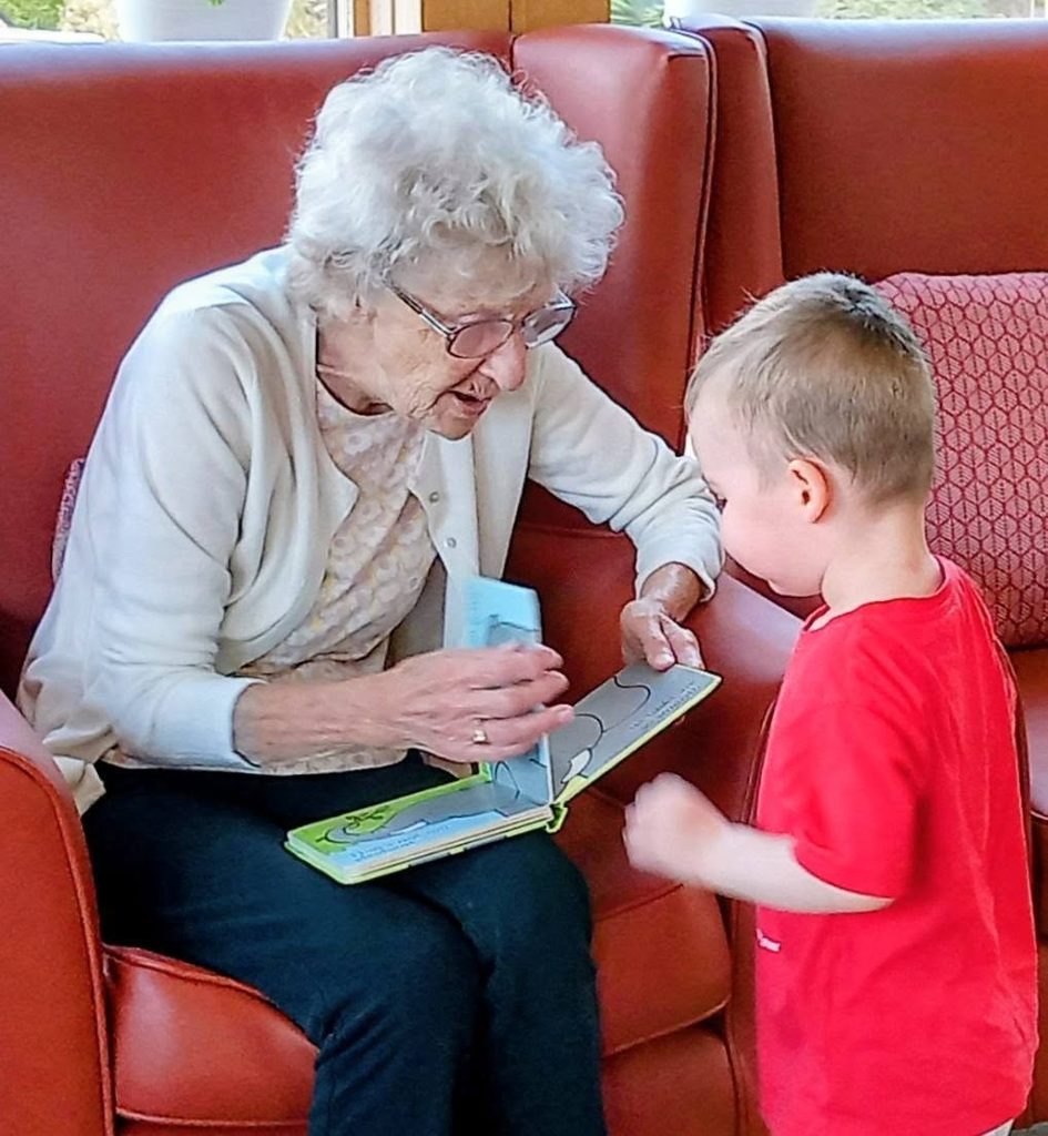 An elderly woman sits on a red chair at Lear House, showing a book to a young boy in Carrie’s Tiney Home Nursery. They interact warmly in a brightly lit room with large windows.