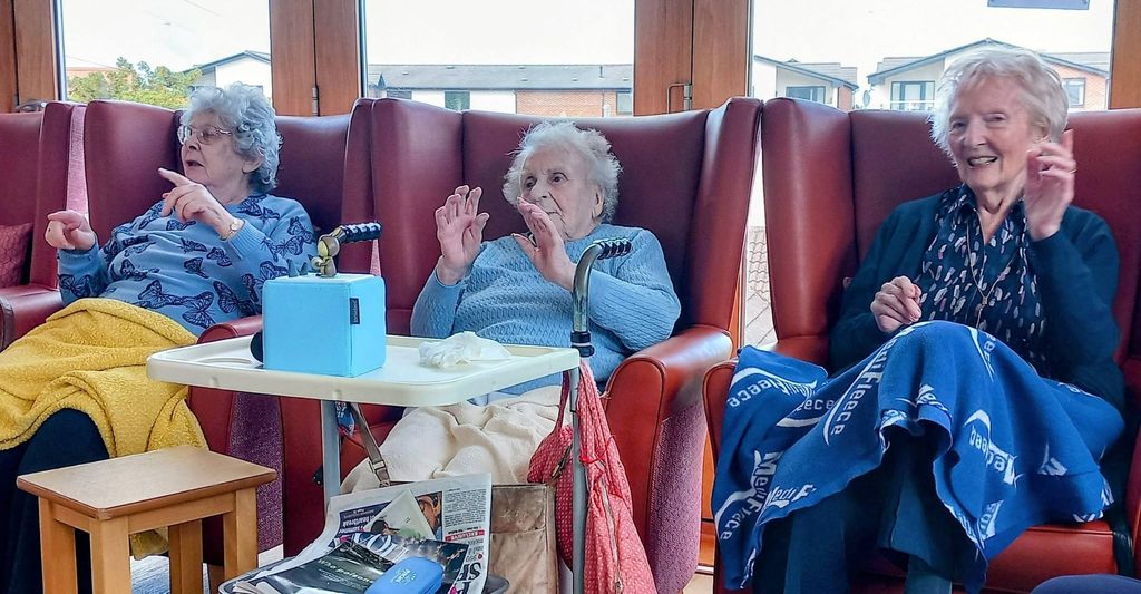 Three elderly women at Lear House sit in red chairs, smiling and gesturing. Zimmer frames are nearby, and blankets cover their laps. A table with a box of tissues and newspapers is set in front of them.