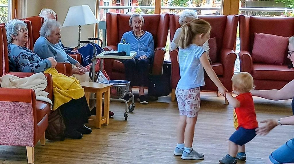 At Lear House, children from Carrie’s Tiney Home Nursery dance hand in hand whilst elderly residents watch from armchairs, fostering a cosy and communal atmosphere.
