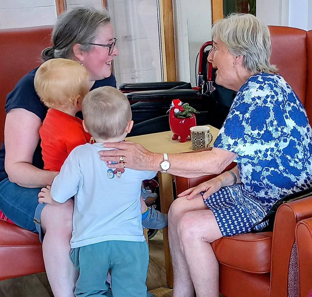 Two women at Carrie's Tiney sit in armchairs chatting whilst two young children stand between them. A small table with a mug and red vase decorates the cosy home nursery setting.