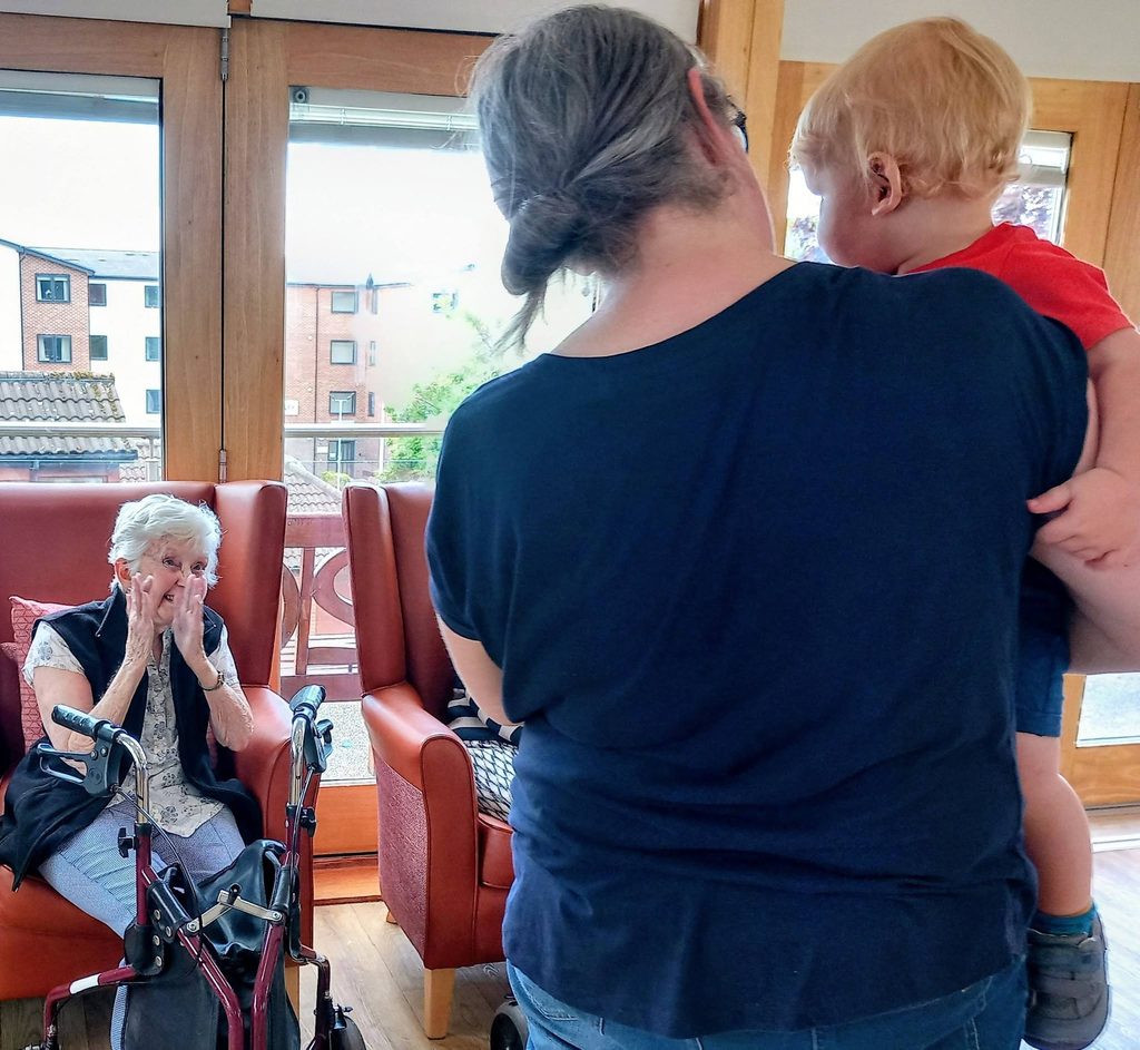 An elderly woman sits in a chair with a Zimmer frame beside her, while a woman holding a young child—possibly from Tiney Home Nursery—stands nearby. Large windows and city buildings are visible in the background.