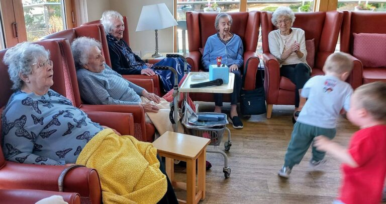 Five elderly women sit in armchairs in a bright room, watching two young children run past. The women look relaxed, with a table holding tissues and a lamp nearby.