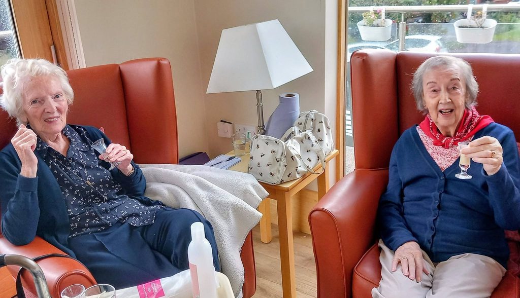 Two elderly women sit in armchairs, smiling and holding drinks. A side table with bags, a lamp, and household items is between them. They are in a cosy, well-lit room with potted plants visible at the window.