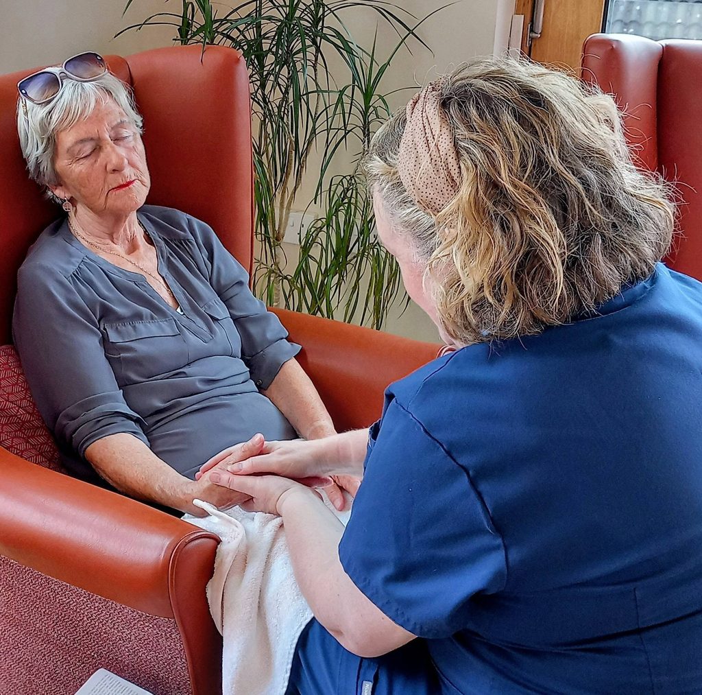 A carer in blue scrubs holds the hands of an elderly woman seated in a red armchair, eyes closed, with a towel on her lap in a well-lit room with a plant in the background.