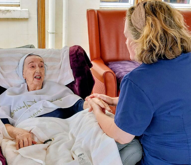 An elderly woman lies in a recliner, wrapped in a blanket, enjoying relaxing afternoons as she holds hands and chats with Jess, her attentive companion seated next to her.