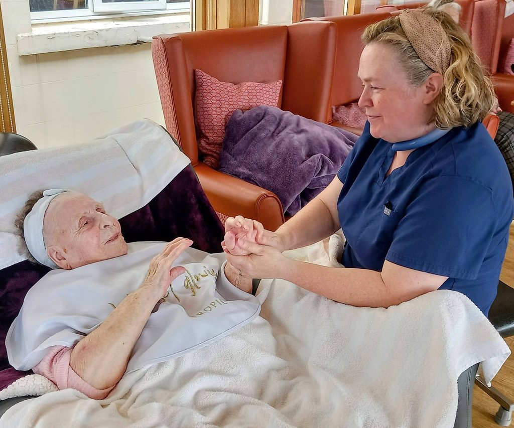 Jess, a carer in navy scrubs, gently holds the hands of an elderly woman during their Pamper Afternoons, both surrounded by blankets and cushions in a cosy room.