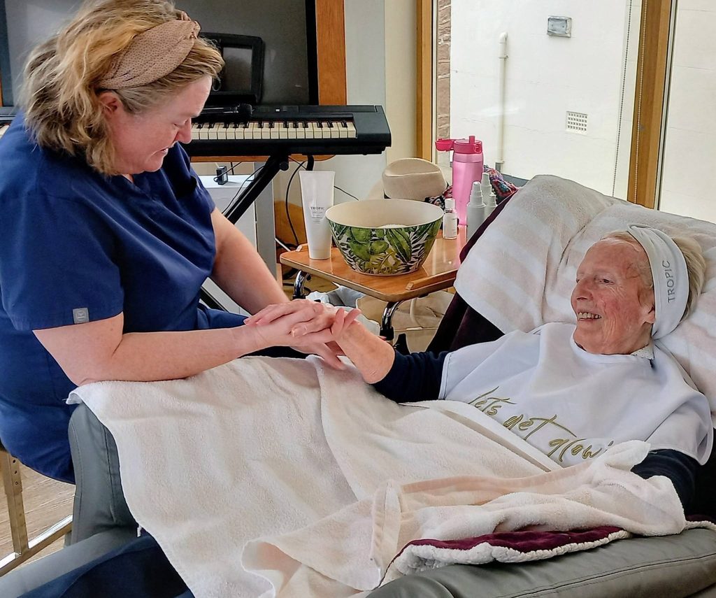 Jess, a carer in navy scrubs, holds the hand of an elderly woman enjoying Pamper Afternoons—she smiles, reclined in a chair with a towel, receiving a manicure in a bright, airy room.