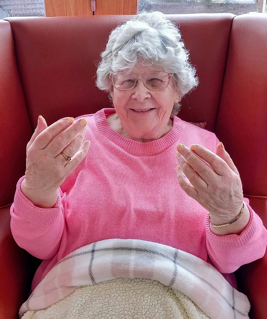Jess enjoys Pamper Afternoons as she sits in a red chair, wearing a pink jumper and blanket, smiling warmly whilst showing her fingernails to the camera.