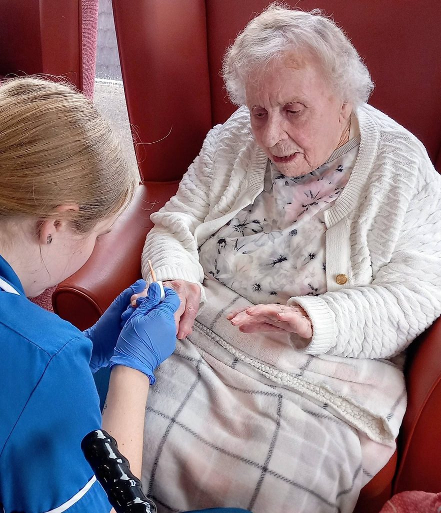 Nurse Jess, wearing blue gloves, examines the hand of an elderly lady enjoying one of our Pamper Afternoons, cosy under a blanket in her chair.