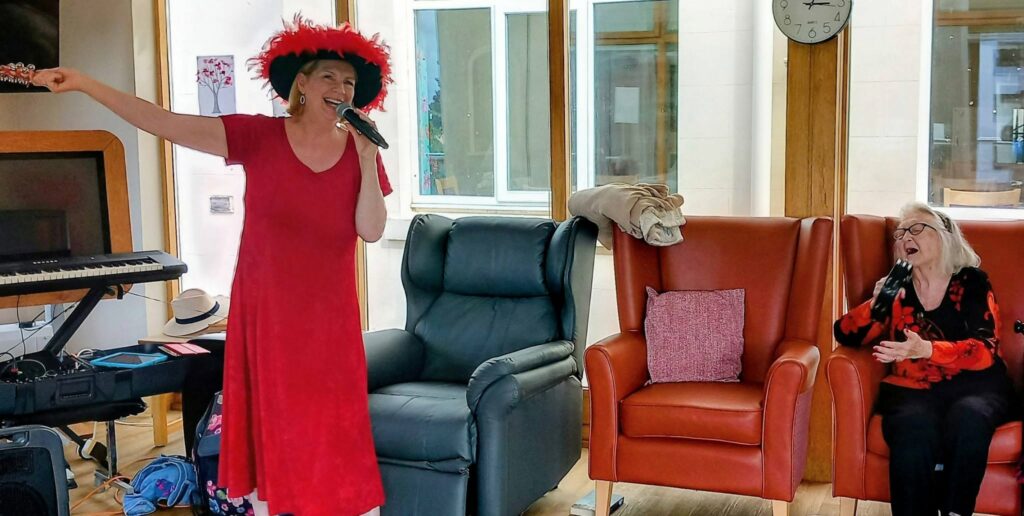 A woman in a red dress and feathered hat sings into a microphone by the armchairs, as another woman in a floral blouse sits and joins her, filling the light-filled room with music.