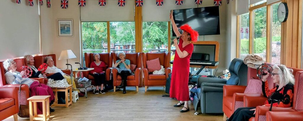 A group of elderly people sit on orange chairs in a festively adorned room as someone in a red dress and feathered hat entertains by singing, Union Jack flags above and the lively spirit of music all around.