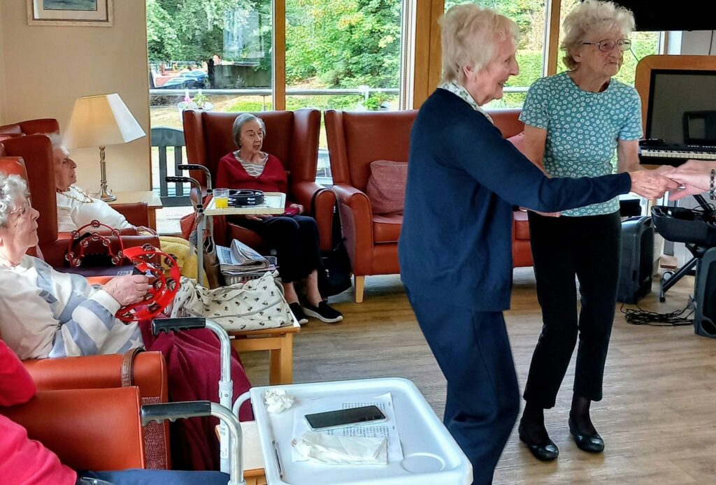 Two elderly women dance joyfully to music in a sunlit communal lounge, whilst three others watch from armchairs. A walking frame and a tray sit in the foreground, completing the lively, heart-warming scene.