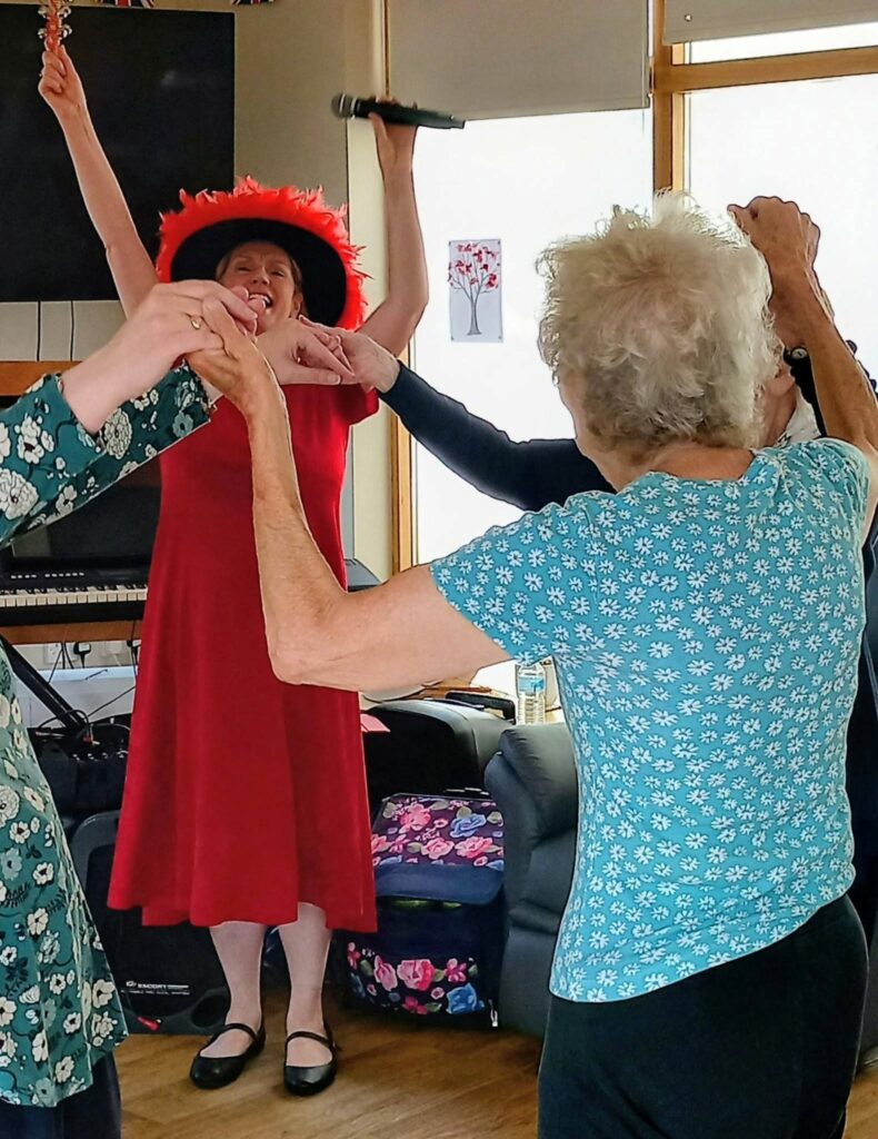 A woman in a red dress and hat sings into a microphone as lively music fills the air, whilst a group of older adults hold hands and dance together in a bright indoor room.