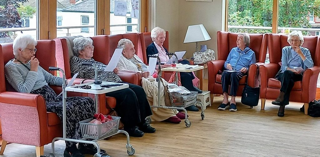Six elderly women sit in armchairs arranged in a semicircle, enjoying friendly conversation and reading together, surrounded by walking frames and side tables in a bright, spacious room.
