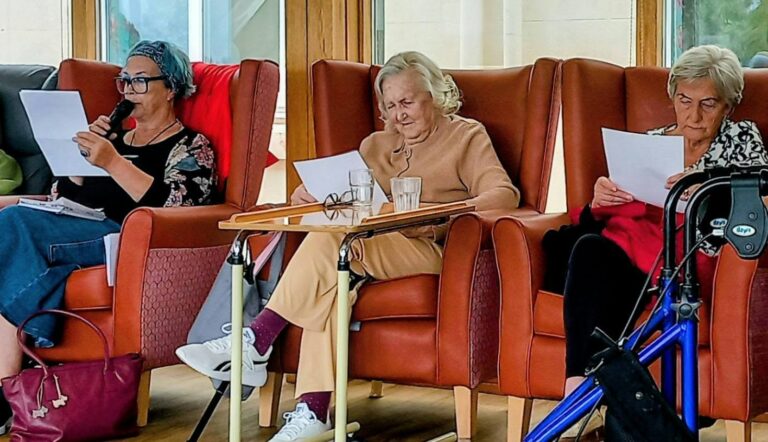 Three elderly women sit in armchairs enjoying a shared reading; one speaks into a microphone, another sips a drink from a tray table, with a walking frame placed nearby.