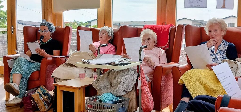 Four elderly women sit in armchairs at Lear House, each with a poetry sheet and wearing microphone headsets. Knitting items and papers suggest a cosy shared reading session in the sunlit room with large windows.