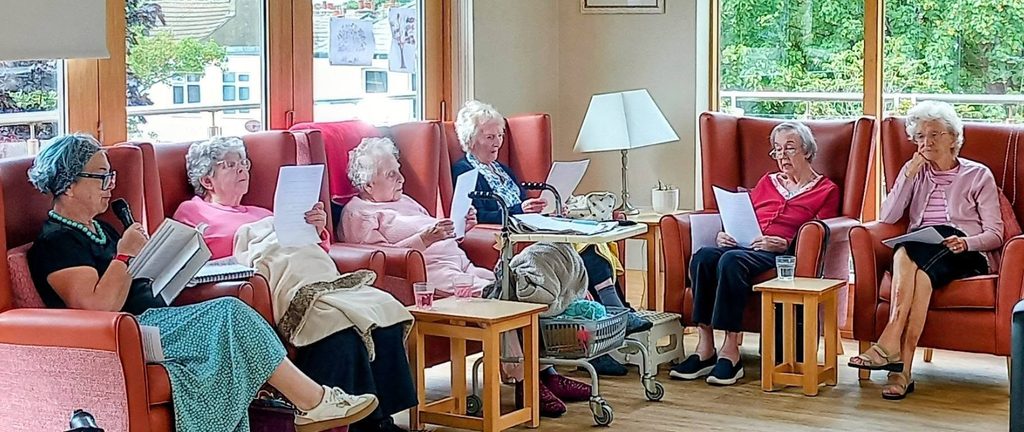 Six elderly women at Lear House share poetry and reading in a bright lounge, seated in a semicircle. Greenery is visible through large windows, and their cosy space exudes warmth and companionship.