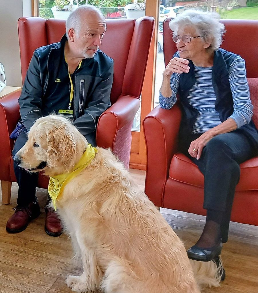 An elderly man and woman sit chatting in red armchairs. Gwyl and Tim, therapy dogs from Therapy Dogs Nationwide, are seated on the floor before them, each wearing a yellow bandana.