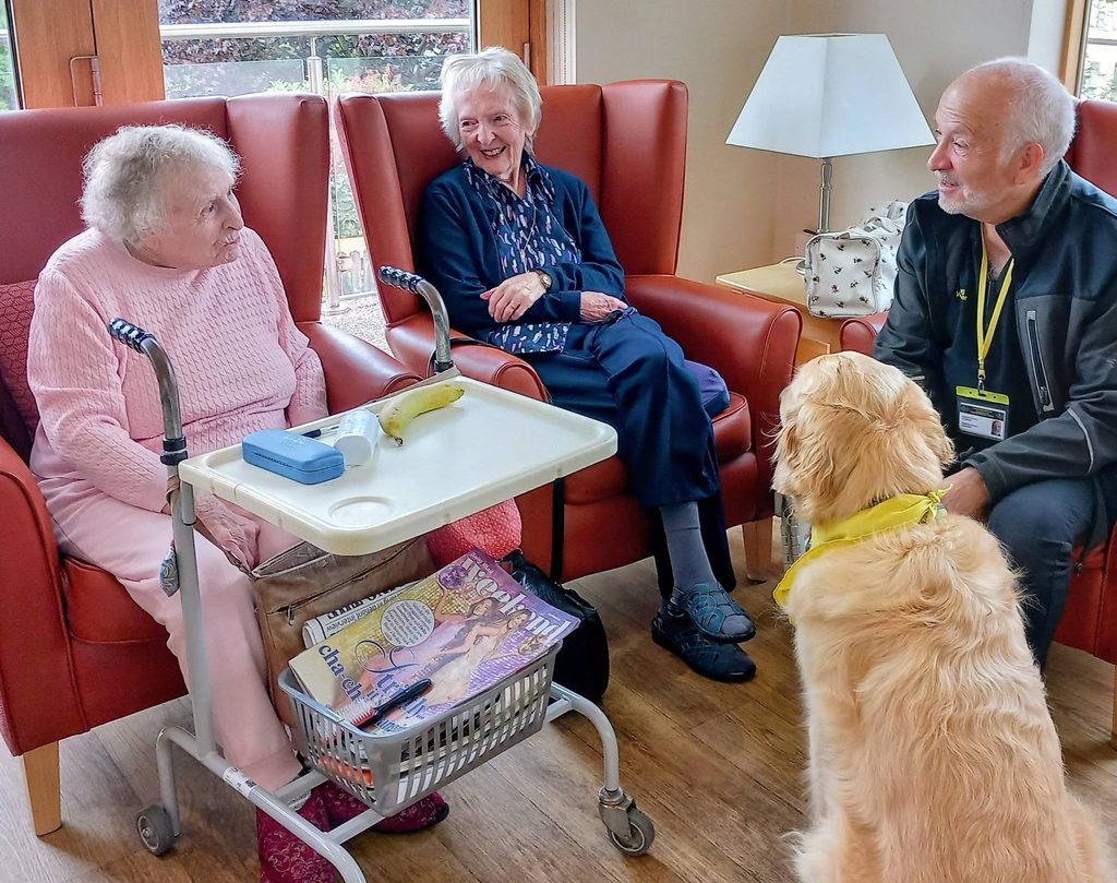 Two elderly women in armchairs chat with a man and his golden retriever from Therapy Dogs Nationwide. Magazines and fruit rest on a tray, creating a cosy care home lounge perfect for “Paws for Thought.”.