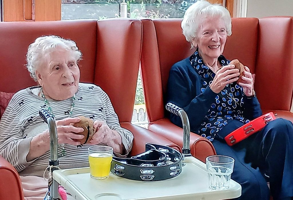 Two elderly women sit in armchairs, smiling and holding sandwiches. A table holds drinks and a walking frame. One woman, with a red tambourine on her lap, laughs as the Two Can Uke music plays quietly in the background.