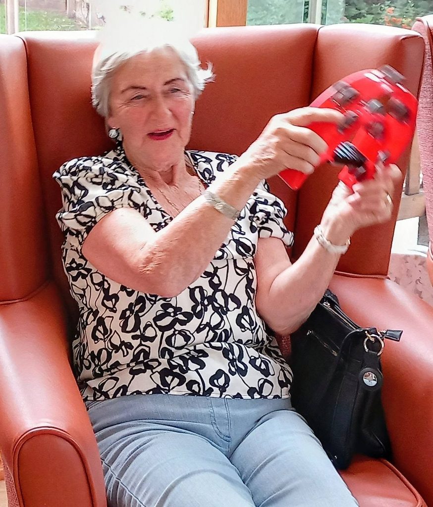 An older woman sits in an armchair, holding a red games controller and appearing engaged. She wears a patterned blouse, light blue trousers, and has a black handbag beside her as Two Can Uke music plays softly in the background.