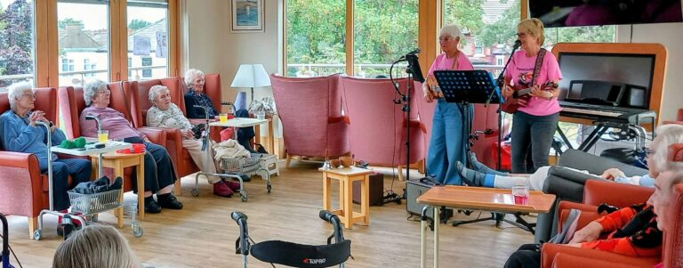 Two women perform music for a group of elderly people seated in a bright communal room with large windows, pink chairs, and walking frames nearby.