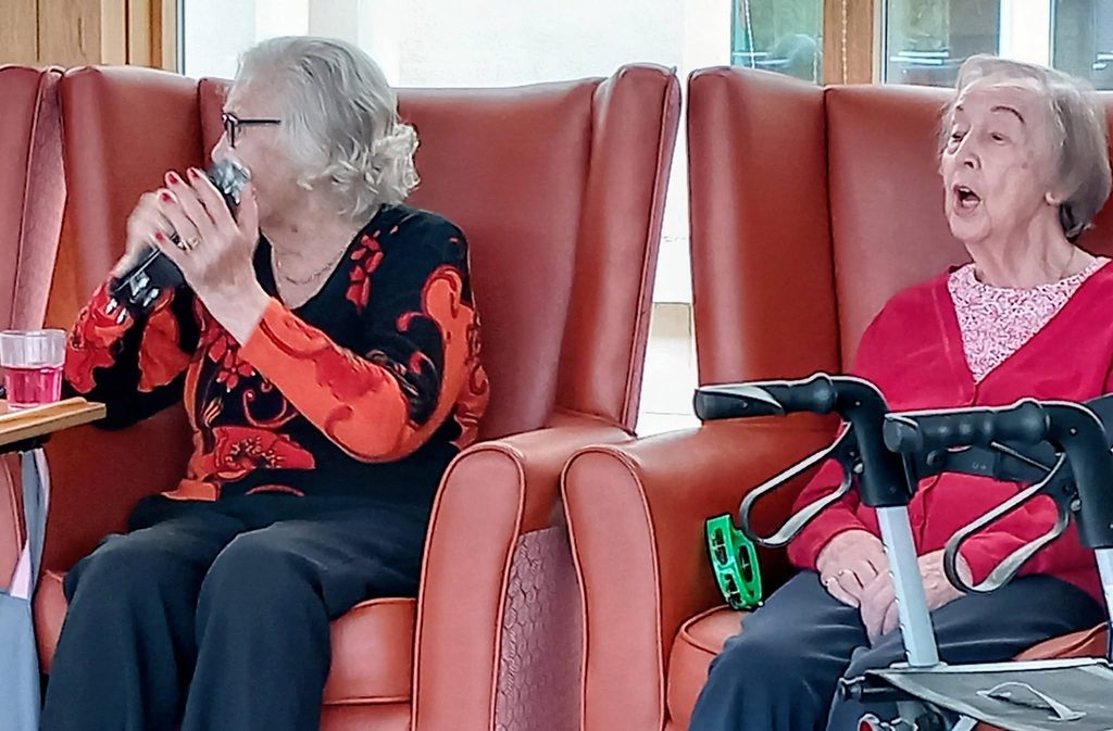 Two Can Uke: Two elderly women sit in armchairs, singing into a microphone together. One has a zimmer frame next to her and a pink drink sits on the table in front of them.
