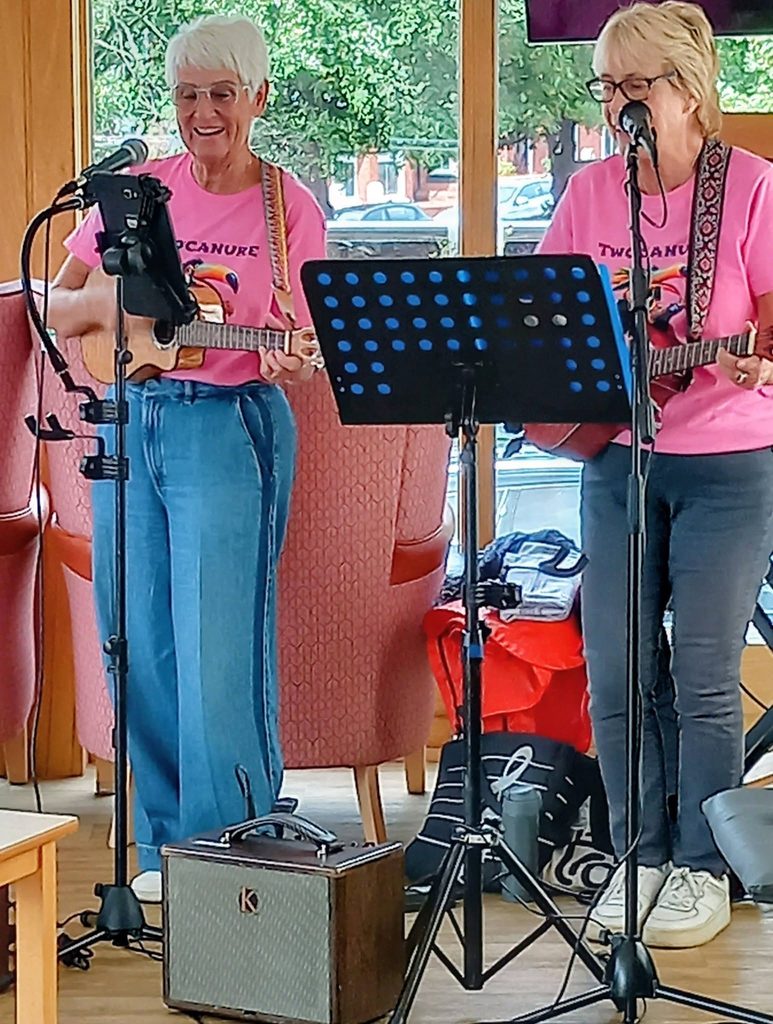 Two Can Uke: Two women in matching pink tops play ukuleles and sing behind a music stand and microphone in a brightly lit indoor space with chairs and large windows.